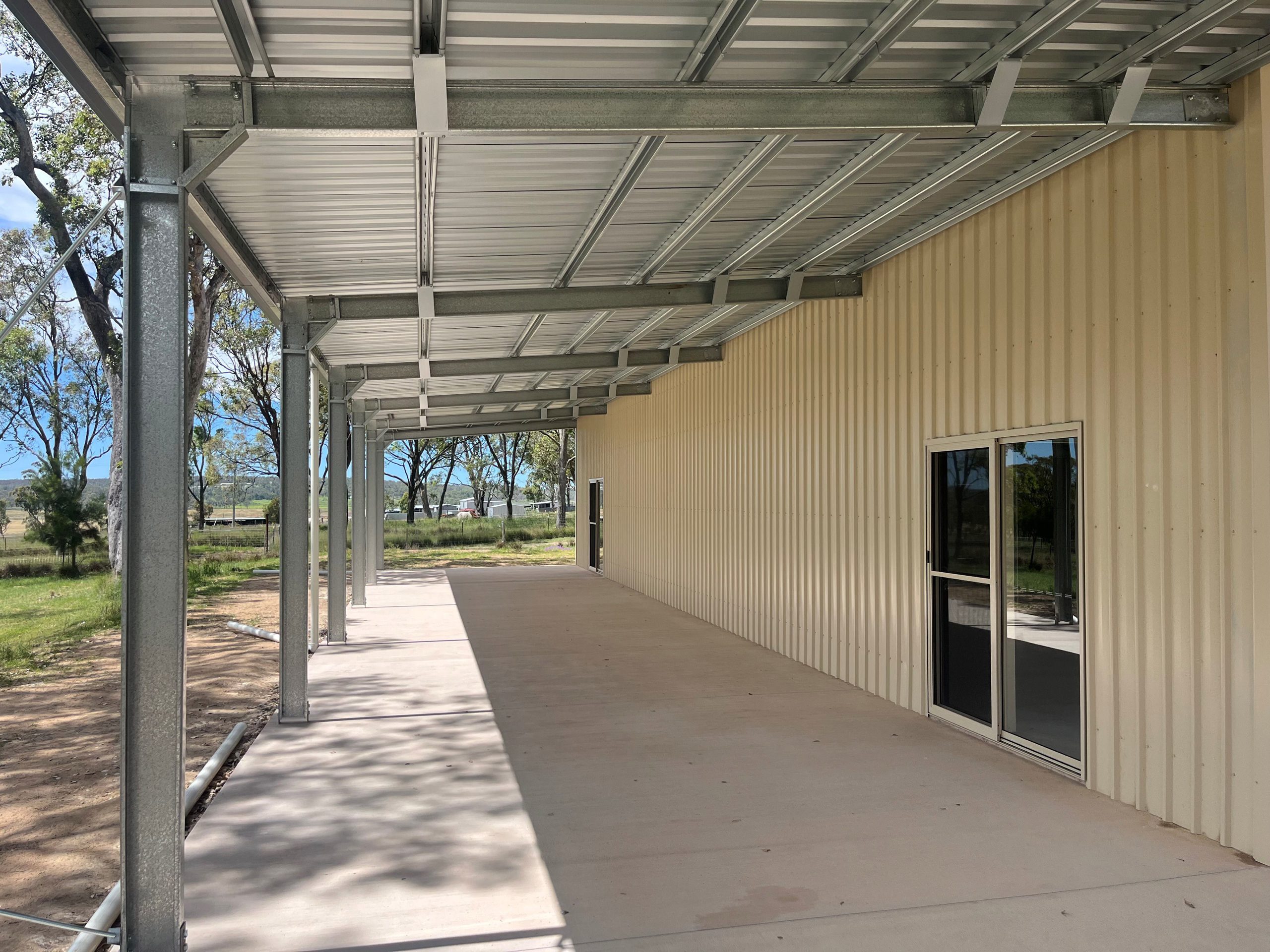 Covered outdoor veranda with steel frame and corrugated iron roof alongside a beige building wall, showing sliding glass doors and a view of trees and fields in the background.