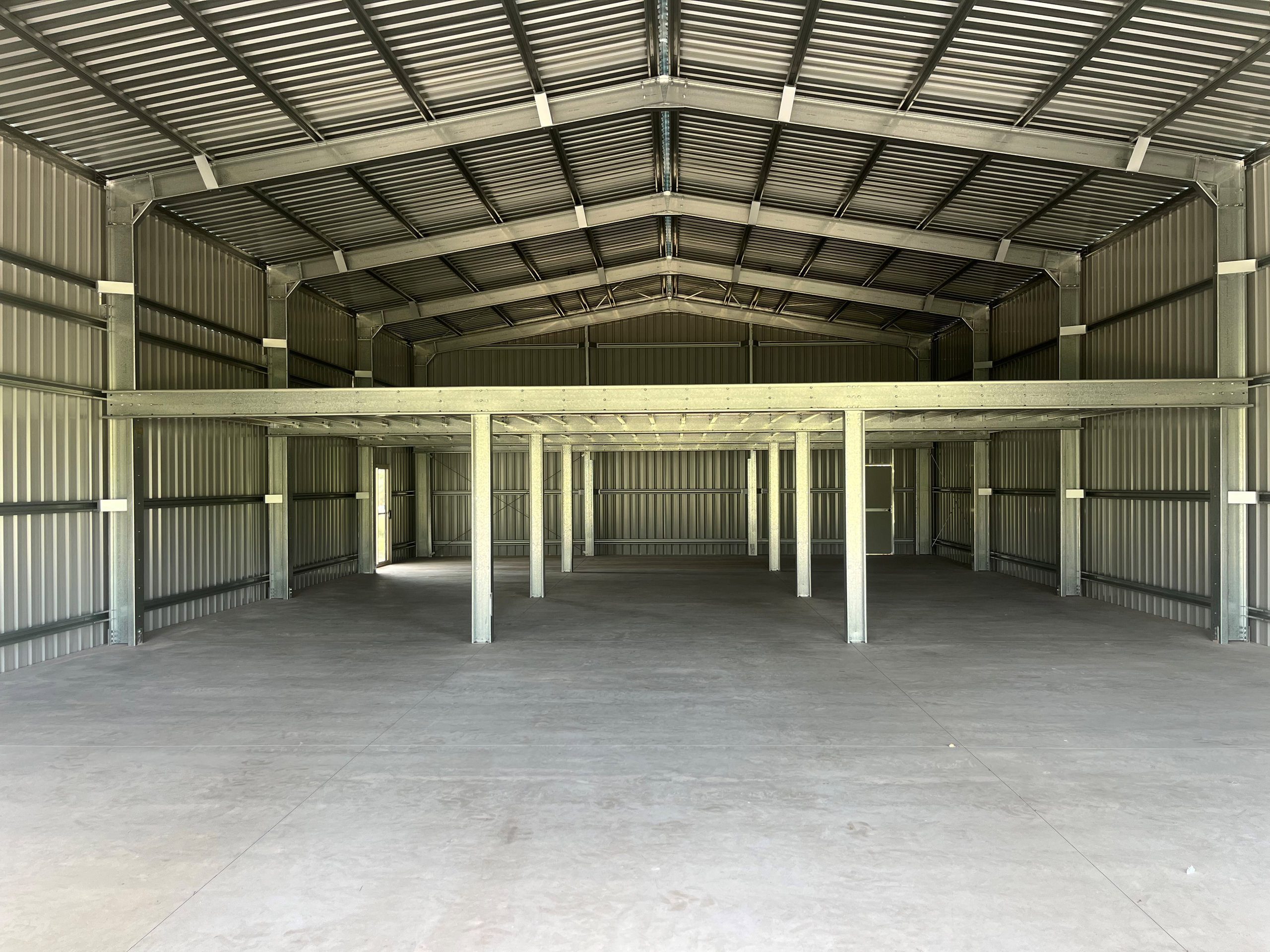 Inside view of a large steel shed with metal walls, roof beams, and a mezzanine supported by steel columns, and a concrete floor, illustrating a solid and spacious storage or workshop area.