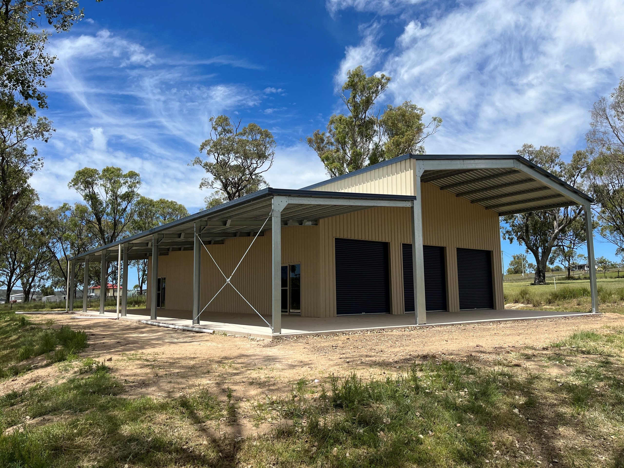 Large beige steel shed with three black roller doors and a long covered verandah area, located in a rural setting with trees and grass under a partly cloudy blue sky.