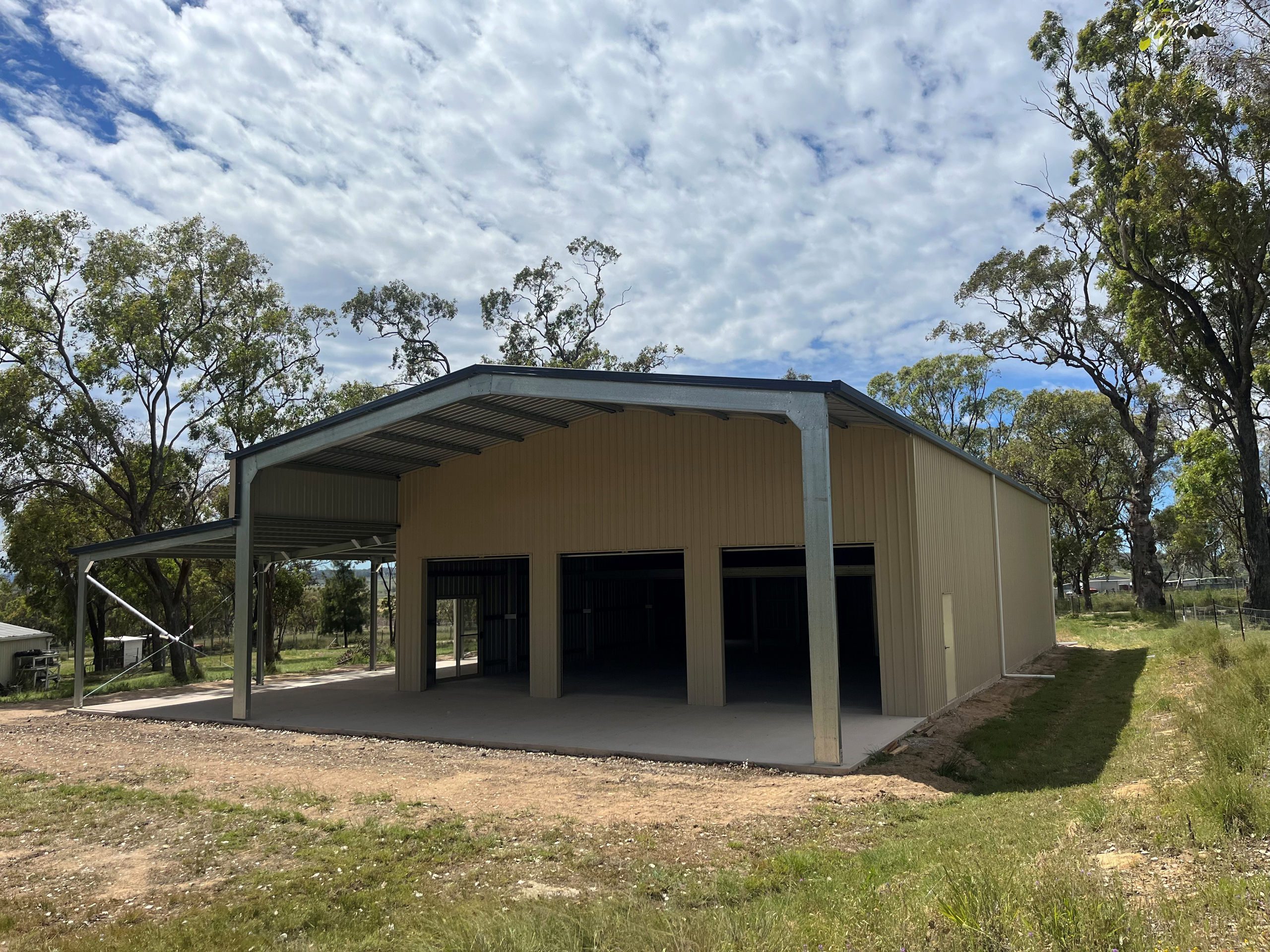New steel shed with beige metal panels and a large open front in a rural setting surrounded by trees and grass under a blue sky with clouds.