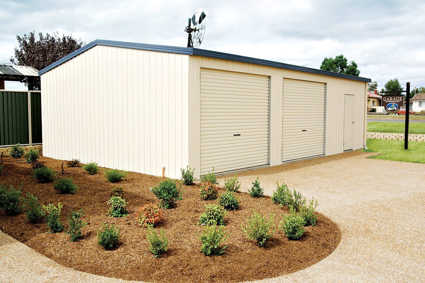 Steel garage with cream-coloured cladding, two closed roller doors and a side door, surrounded by freshly mulched garden beds with small green shrubs and a gravel driveway under a cloudy sky.