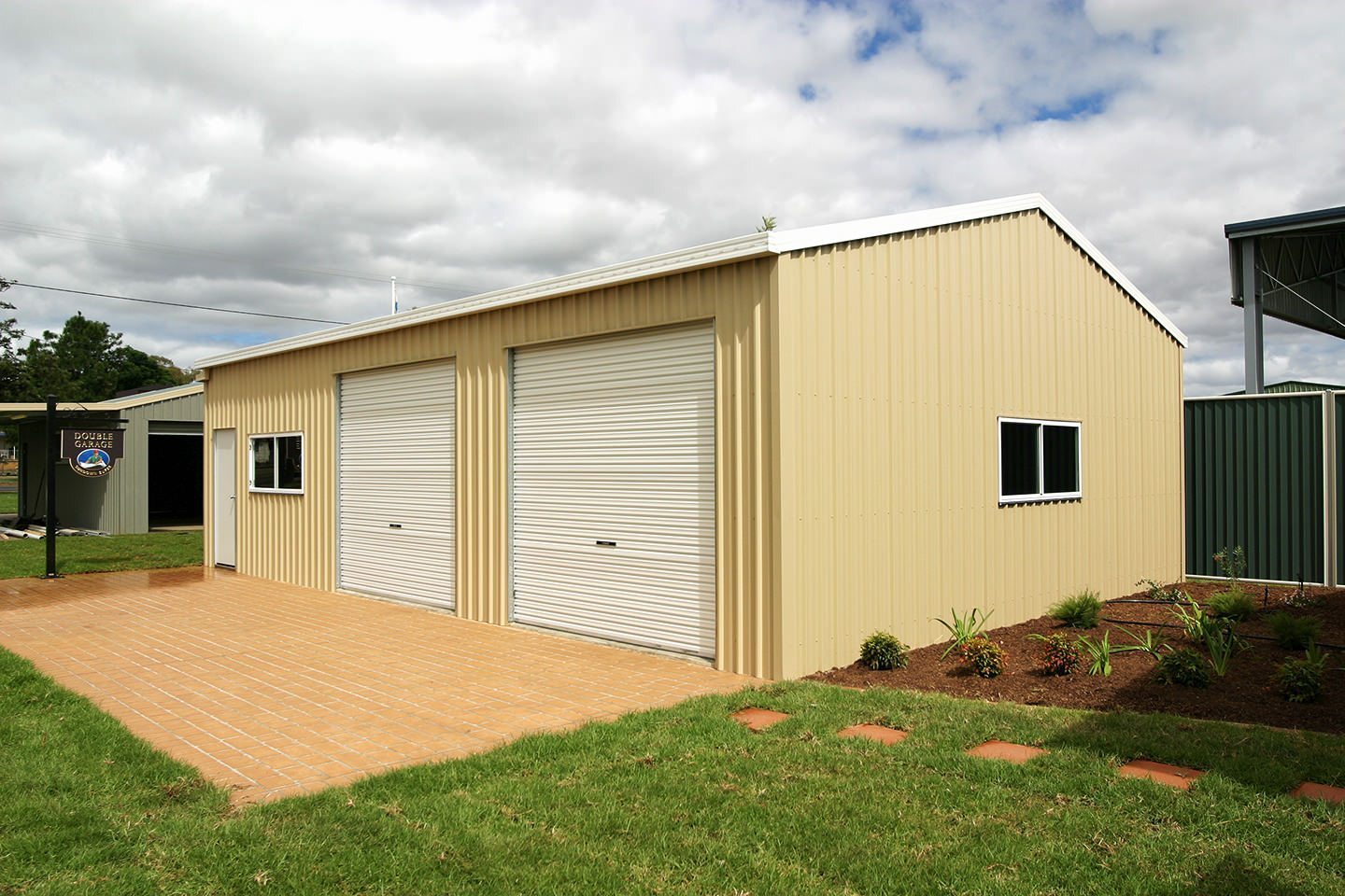 Beige double garage with two white roller doors, paved driveway in front, small windows on the side, and a surrounding green lawn with garden beds.